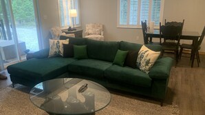 Een woonkamer met een groene modulaire bank, een glazen salontafel en een eethoek met een houten tafel en stoelen.