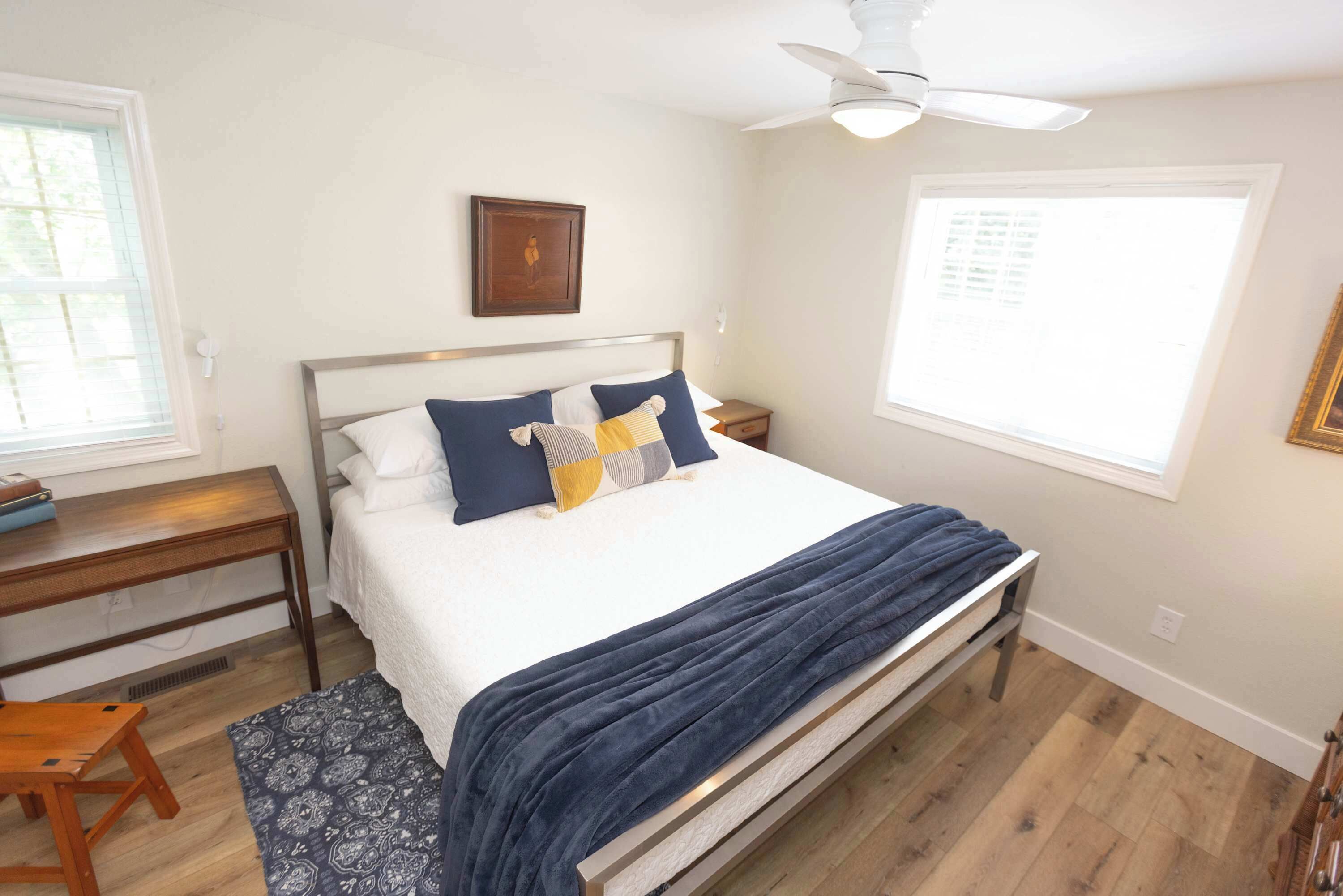 A bedroom with a bed, a desk, a chair, a ceiling fan, and a window.