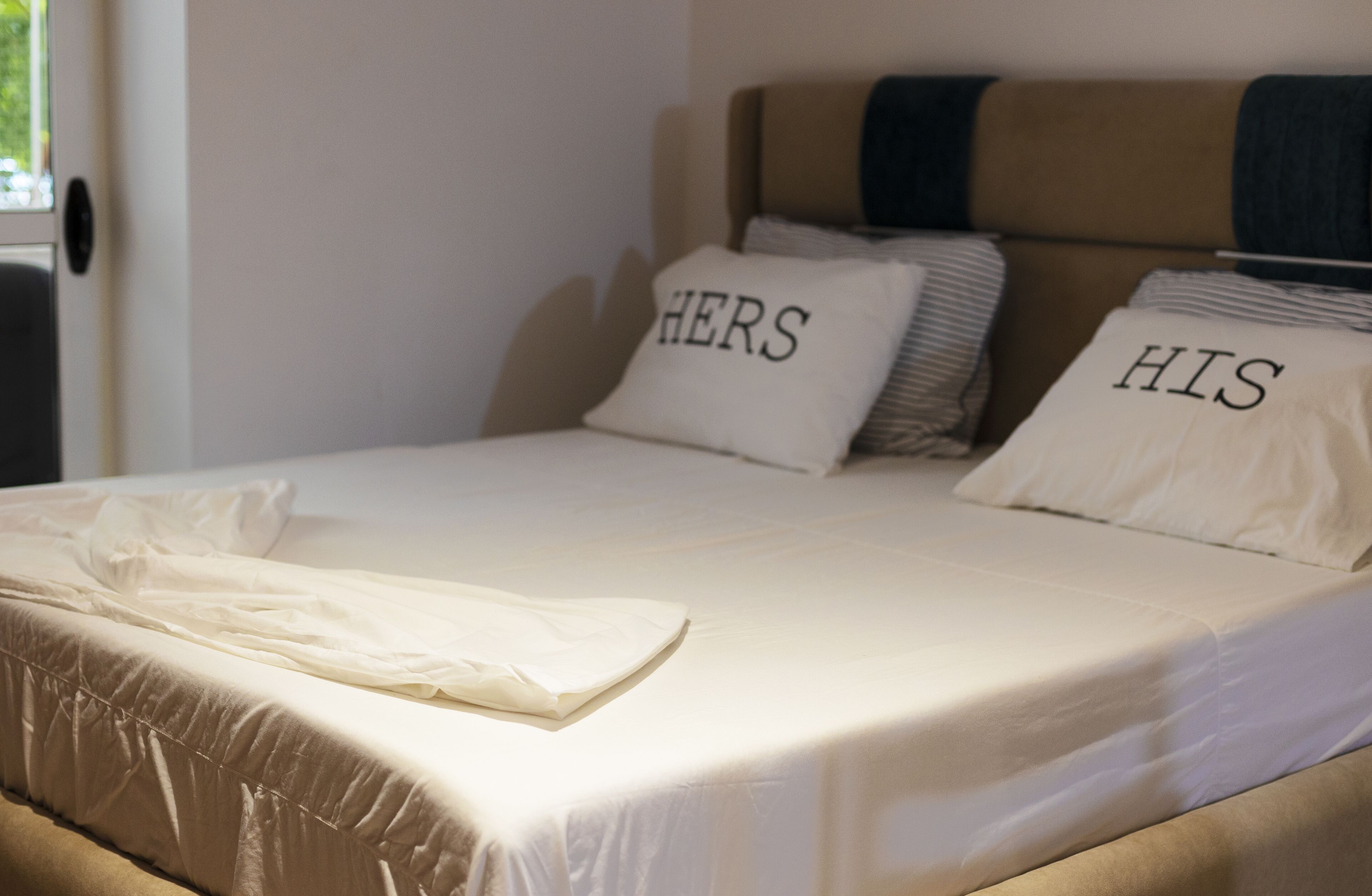 A neatly made bed with pillows labeled "HERS" and "HIS" in a room with a beige headboard and a window.