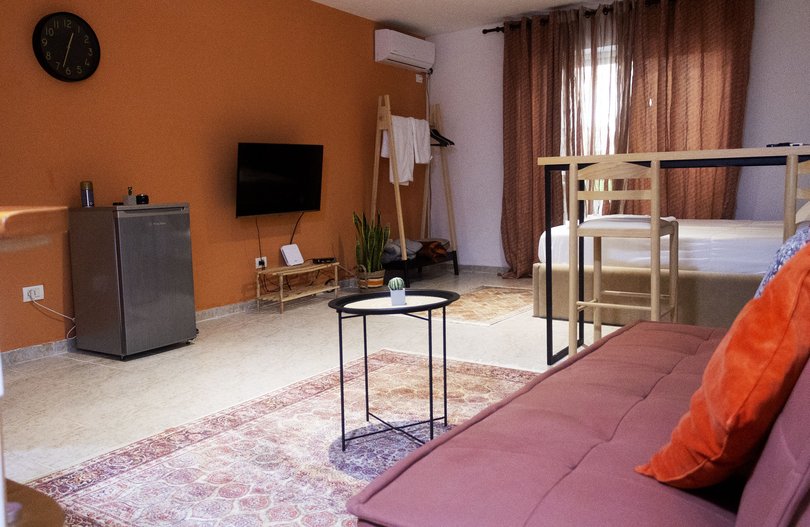 A compact hotel room with a sofa, a small table, a TV, a bed with a wooden frame, a wardrobe, and a plant.