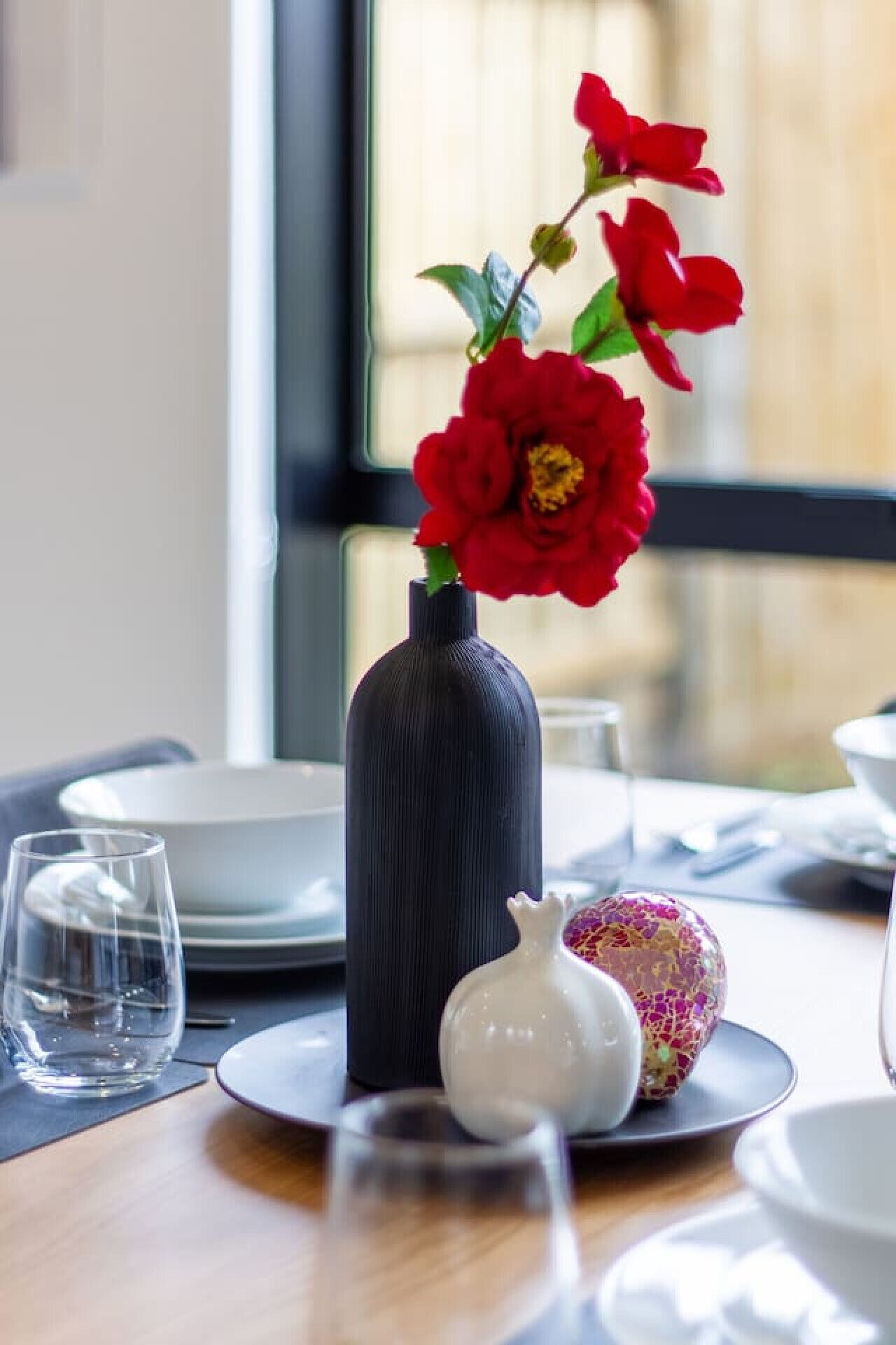 Una mesa de comedor con un jarrón de flores rojas, un huevo decorativo y una jarra blanca pequeña.