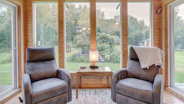 Ein Holzblockhaus-Interieur mit zwei Ledersesseln, einem kleinen Tisch und einer Lampe. Große Fenster bieten Blick auf einen Garten und Bäume.