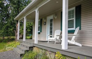 Een overdekte veranda met een houten trap naar een terras, twee witte Adirondack-stoelen, en een huis met groene luiken en een bord met het nummer '11'.
