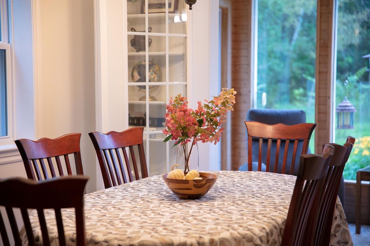 Una mesa de comedor con mantel estampado, rodeada de sillas de madera, y un jarrón de flores sobre la mesa.