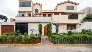 A modern two-story house with a central entrance, a covered driveway, and a small garden area with plants and a fence. - Hermoso y Tranquilo Loft con Netflix Incluido (Lima)