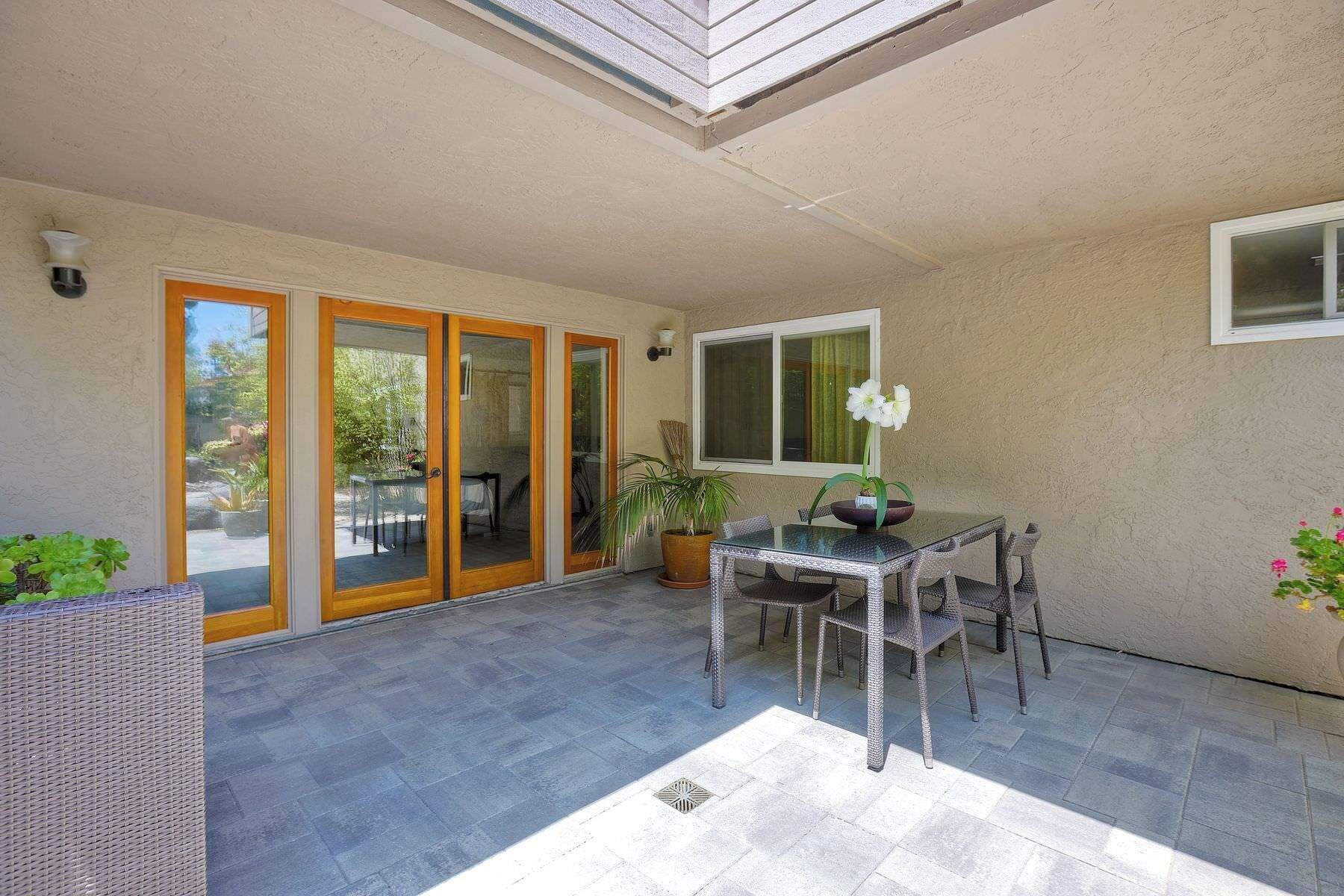 Une terrasse couverte avec une porte vitrée donnant sur un jardin, une table avec des chaises et une plante en pot.