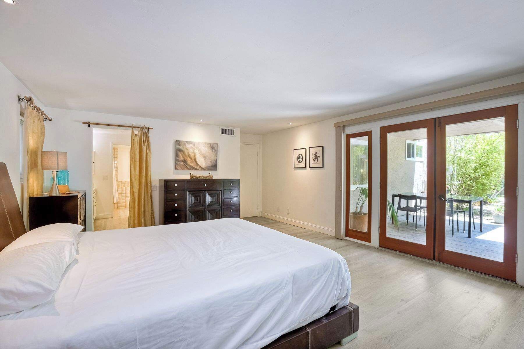 A bedroom with a large bed, a dresser, a lamp, and a view of an outdoor patio.