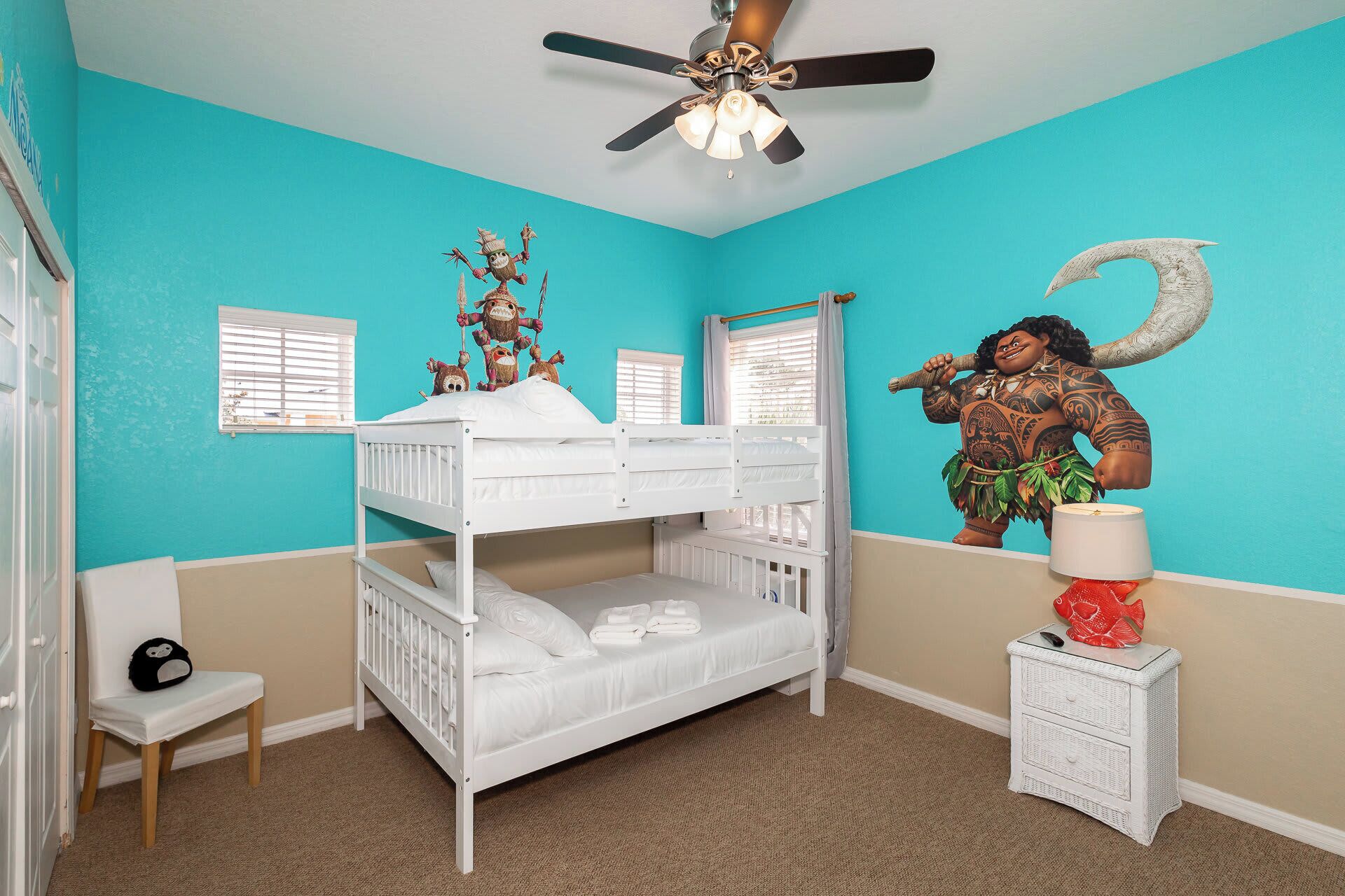 A room with a bunk bed, a ceiling fan, a wall mural, a lamp, and a small table with a decorative item.
