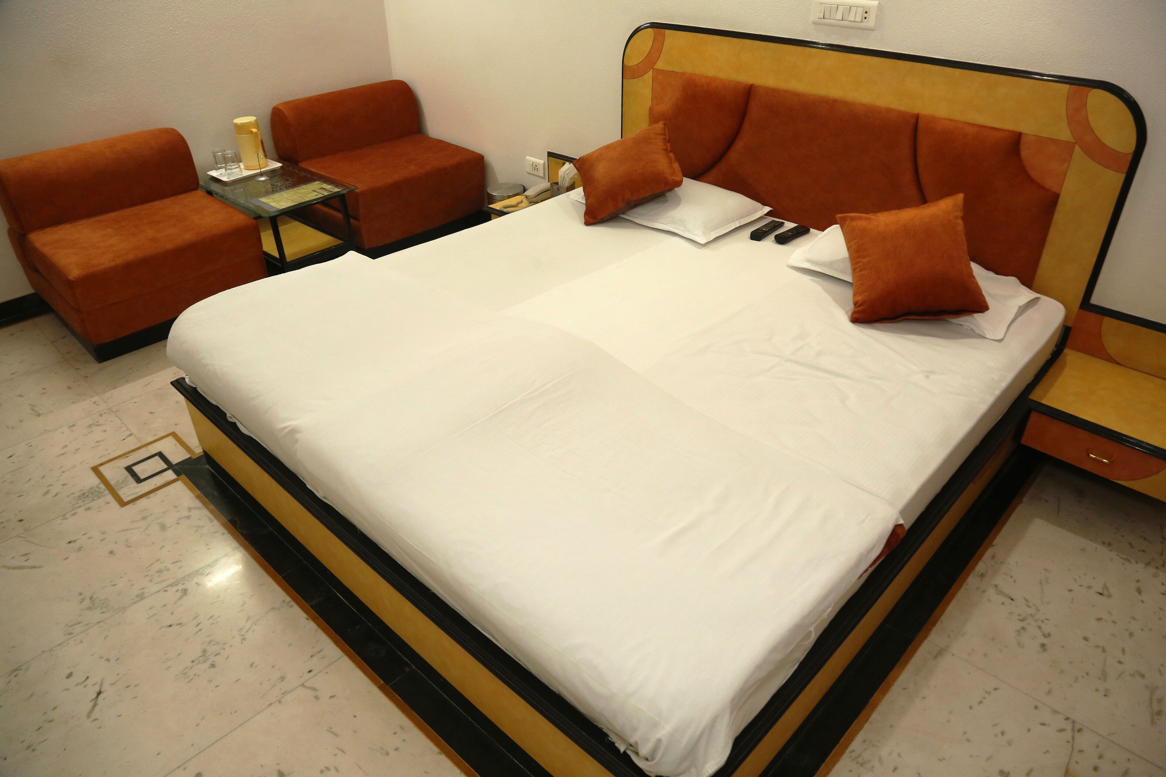 A hotel room with a bed, two orange armchairs, a small table with a glass, and a nightstand.