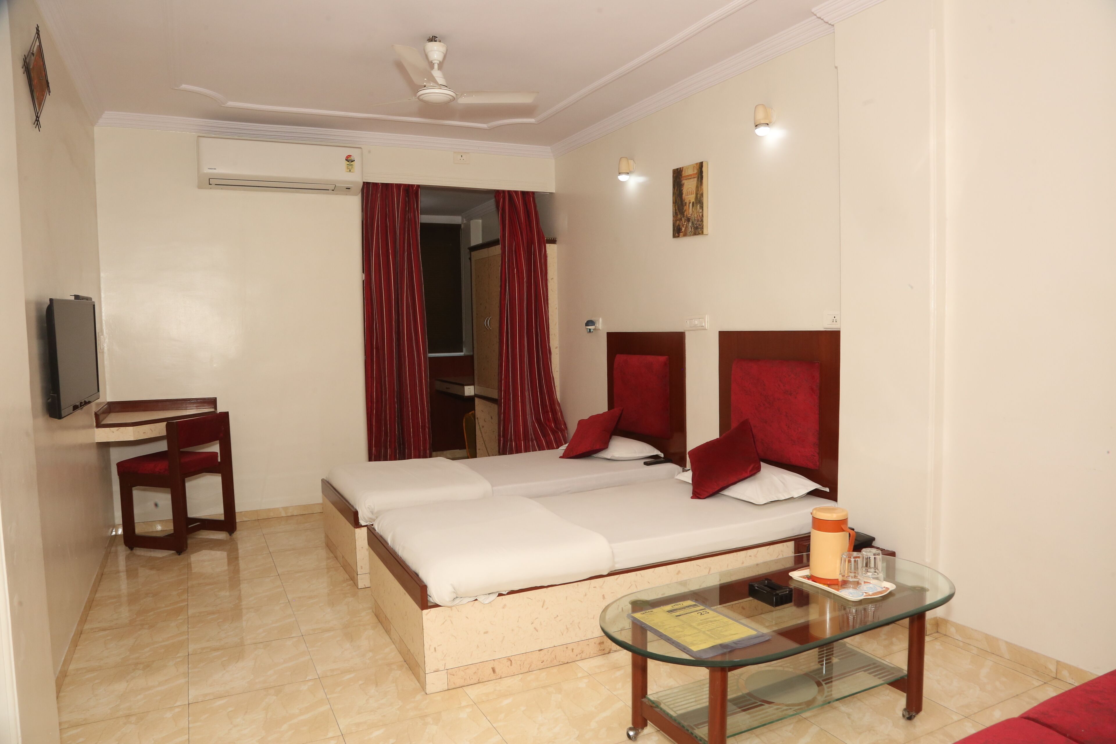 A hotel room with a bed, a desk, a chair, a television, and a glass table with a book and a bottle.