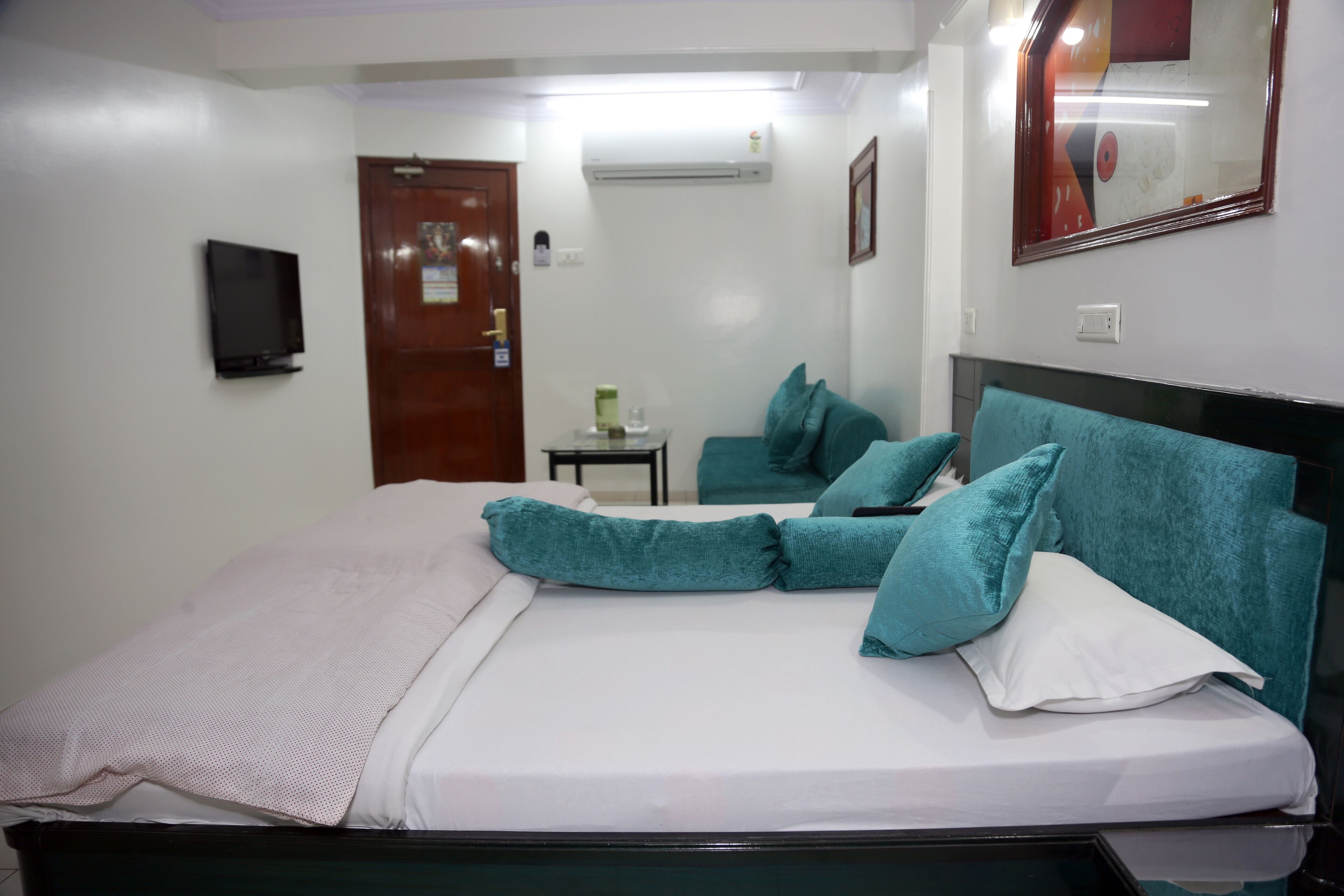 Una cama bien hecha con almohadas verde azulado y blancas, una mesita de noche con una lámpara y un televisor montado en la pared.