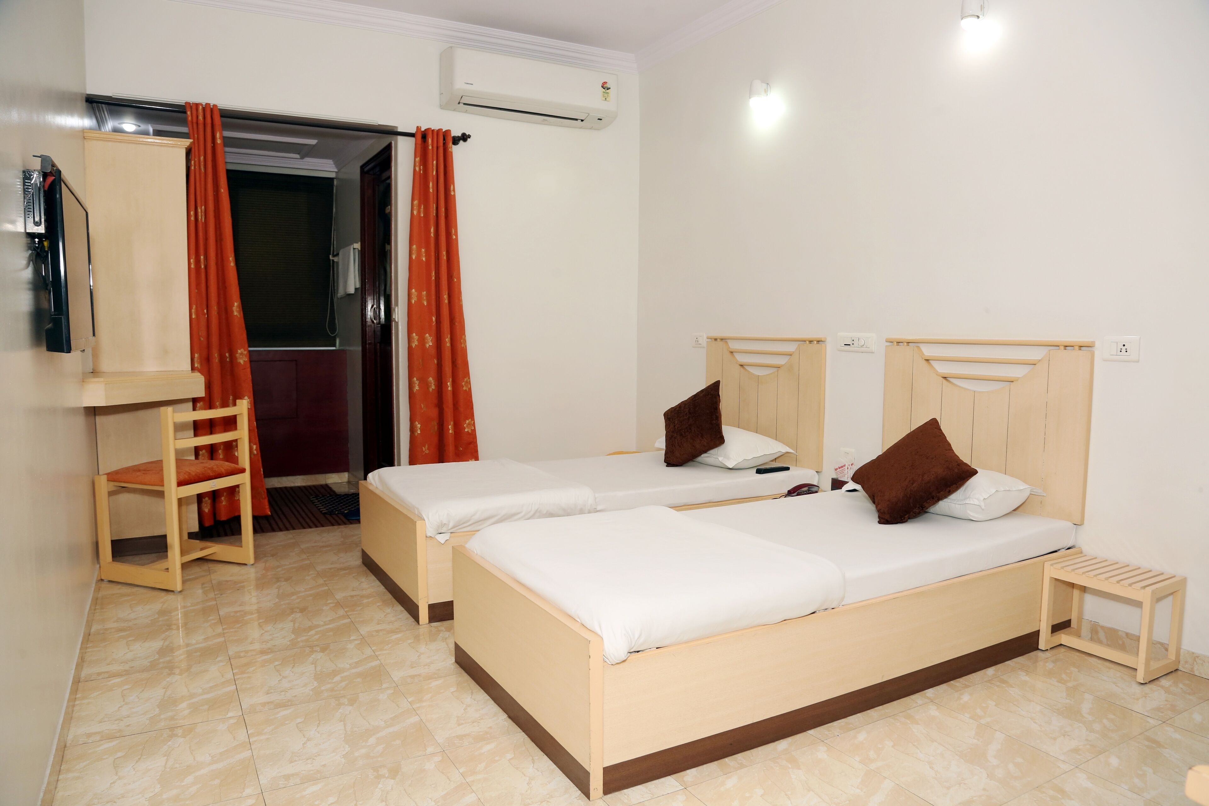 A room with two beds, a wooden chair, a small stool, an air conditioning unit, and a door with red curtains.