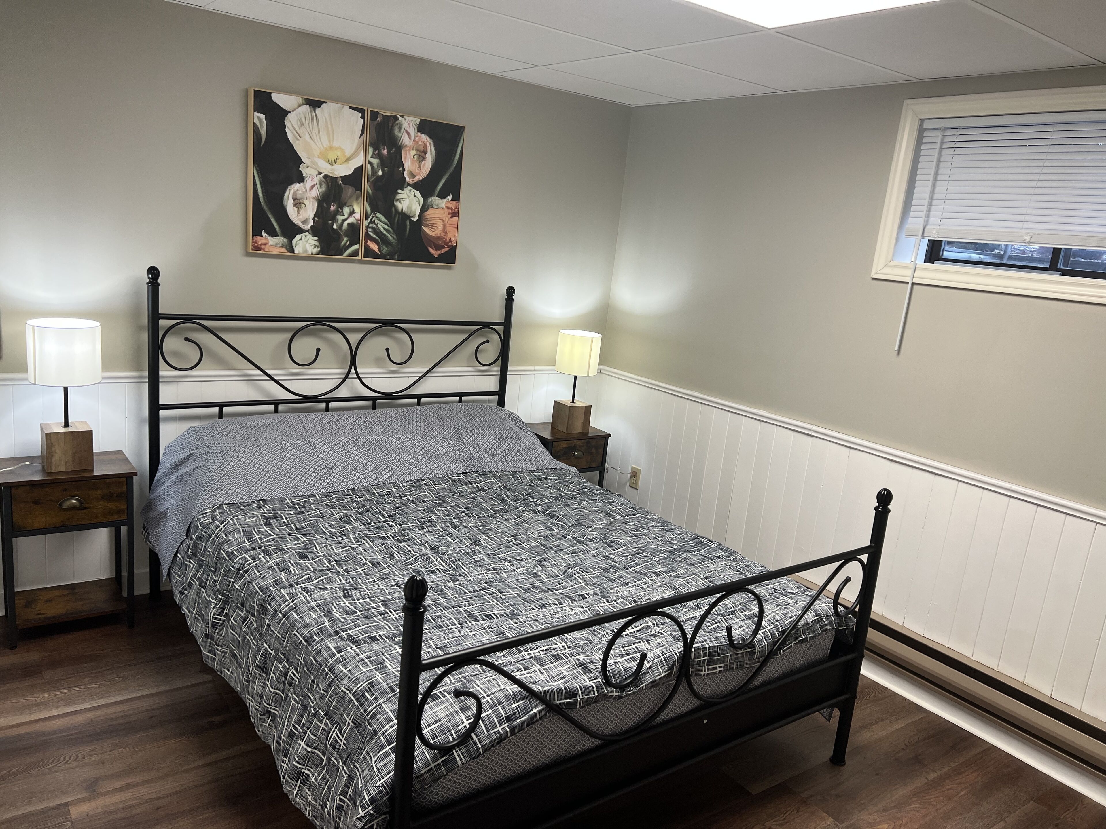 A bedroom with a bed, two bedside tables, a wall-mounted lamp, and a window with blinds.