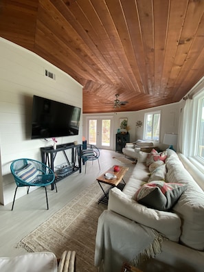 A spacious living room with a wooden ceiling, a flat-screen TV mounted on the wall, a sofa set, and a dining area with chairs and a table.
