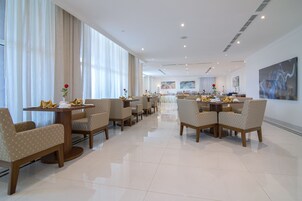 Un amplio comedor de hotel con varias mesas y sillas, ventanas grandes con cortinas y obras de arte modernas en las paredes.