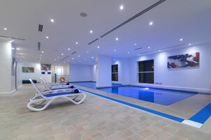 Una piscina interior con azulejos azules, sillones de descanso e iluminación empotrada en el techo.
