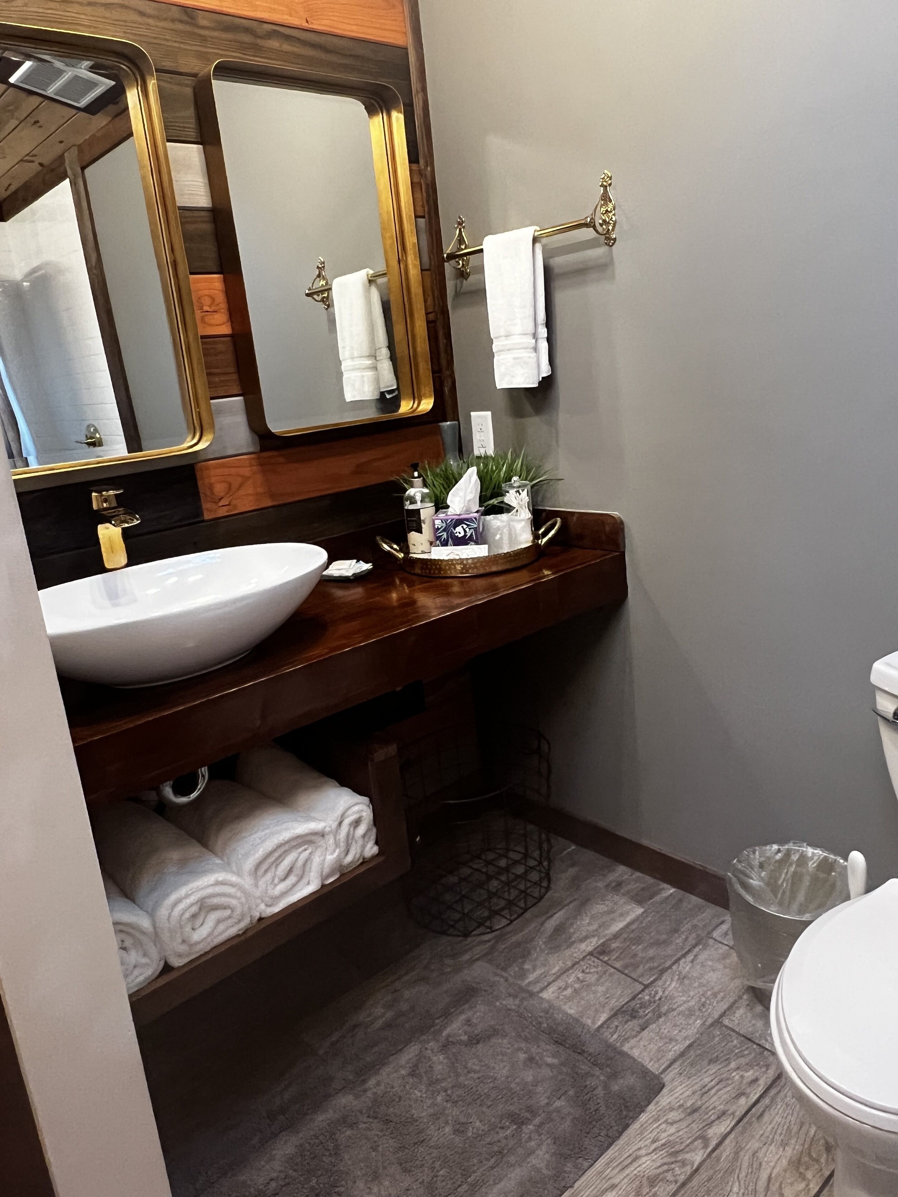 A bathroom with a wooden vanity, a white sink, a large mirror with a gold frame, and a towel rack with white towels.