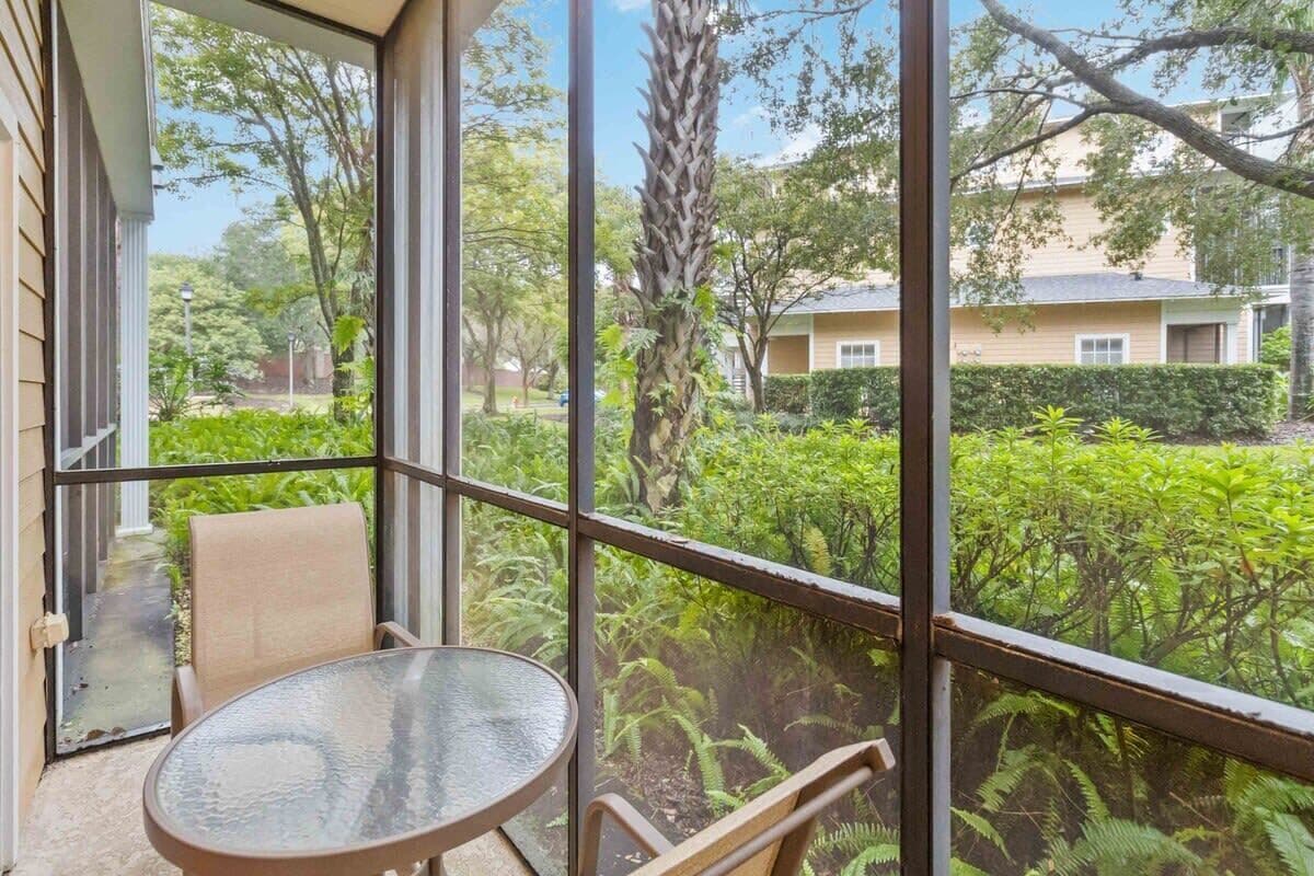 A balcony with a glass table and chairs, overlooking a garden with various plants and a building in the background.