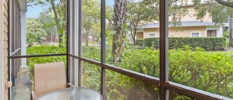 A balcony with a glass table and chairs, overlooking a garden with various plants and a building in the background.