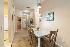 Uma área de refeições com mesa de vidro, cadeiras de madeira, luminárias pendentes, uma pintura na parede e um corredor que dá para uma cozinha.