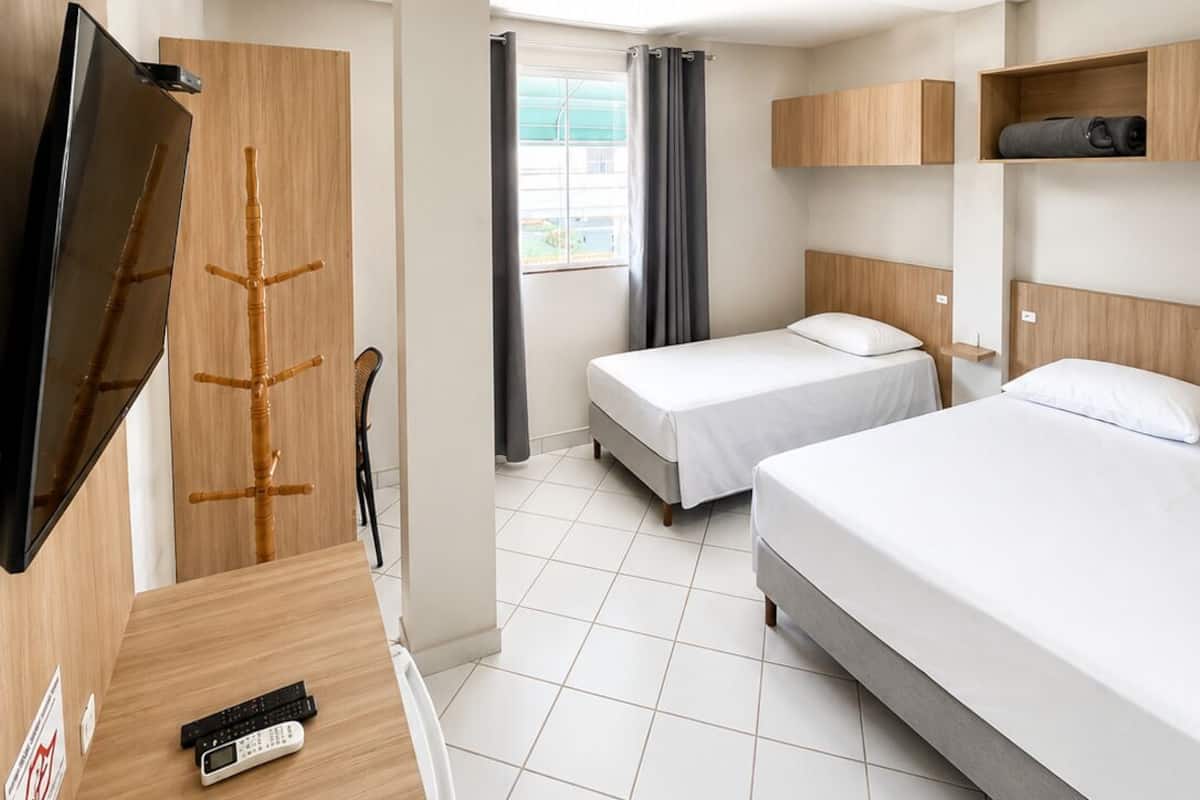 A hotel room with two beds, a TV, a desk with a chair, a window with curtains, and a small shelf with a bag.