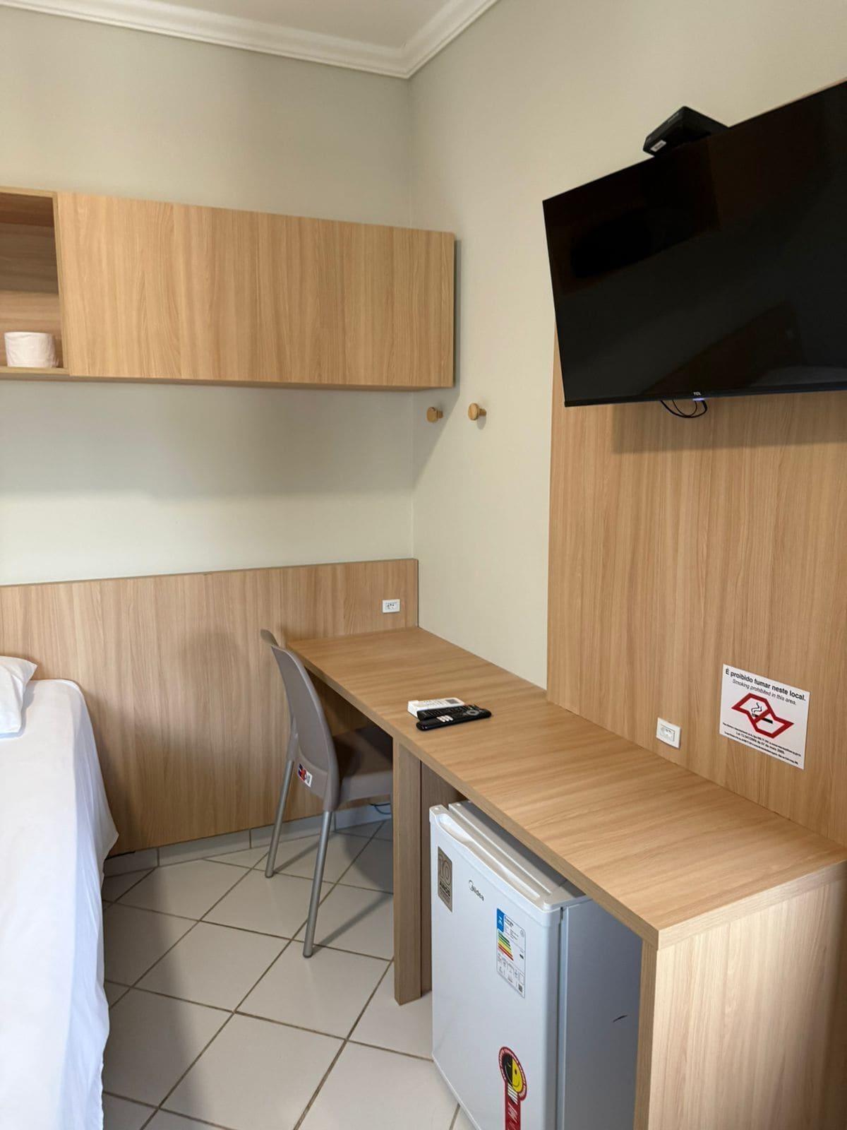 A compact hotel room with a kitchenette, a bed, a desk, a chair, a TV mounted on the wall, and a refrigerator.