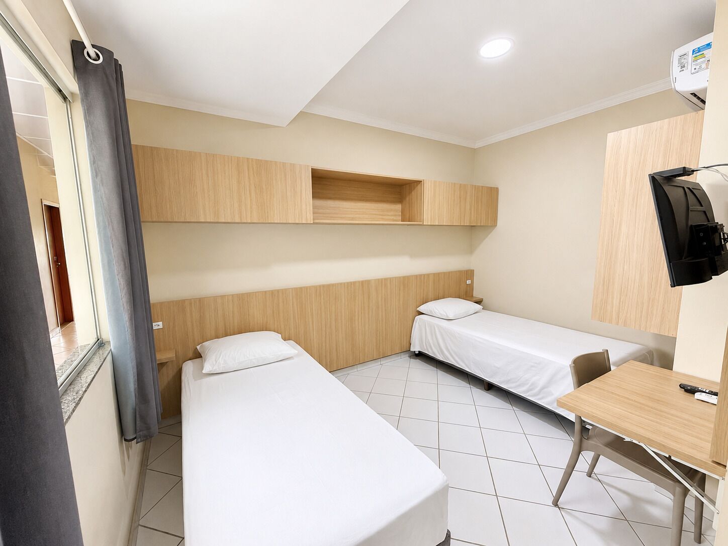 Una habitación de hotel con dos camas individuales, un escritorio, una silla, un televisor y una ventana con cortinas.