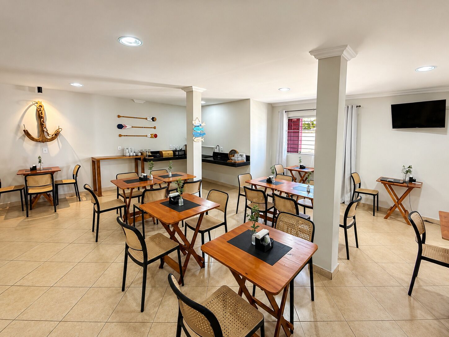 A hotel dining area with multiple tables and chairs, a kitchen in the background, and a TV mounted on the wall.