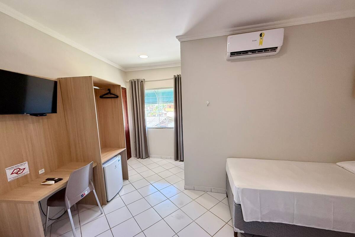 A hotel room with a bed, a desk with a chair, a TV mounted on the wall, and an air conditioning unit.