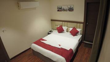 A hotel room with a bed, a nightstand, an air conditioning unit, and a door.