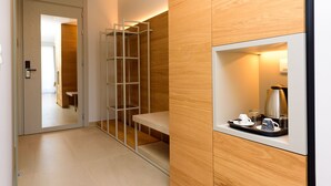 Una habitación de hotel moderna con una pared de madera, un estante empotrado con cafetera, tazas y vasos, y una puerta corredera que da a otra habitación.