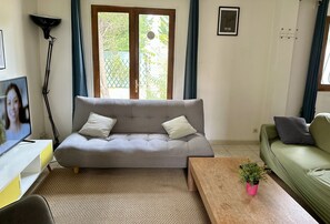 Ein Wohnzimmer mit grauem Sofa, einem Holztischchen mit Pflanze, einem Fernseher auf einem weißen Schrank und einem grünen Sofa im Hintergrund.