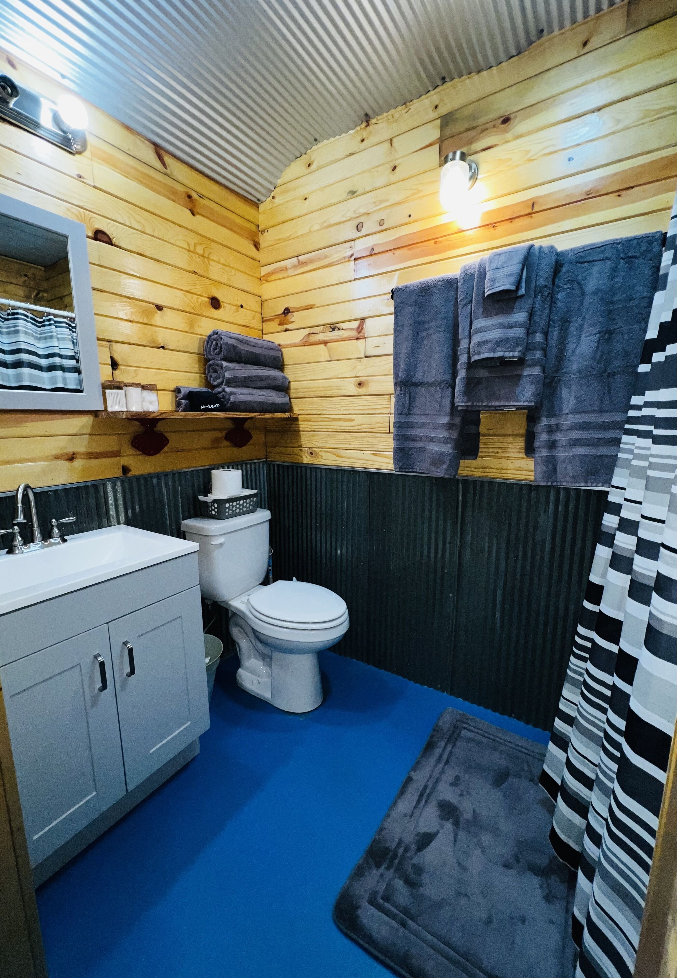 Ein kleines Badezimmer mit Holzwänden, einem weißen Waschbecken, einer Toilette und einem blauen Boden.