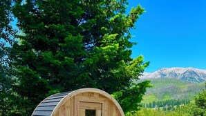 Una construcció de fusta amb forma de bóta i una porta, situada sobre una superfície de formigó, amb una zona verda i arbres al fons.