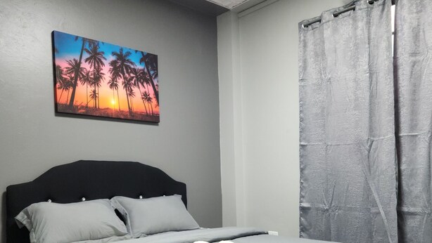 Un lit bien fait, avec du linge de lit gris et une serviette pliée, un tableau représentant des palmiers au coucher du soleil accroché au mur, et un rideau gris.