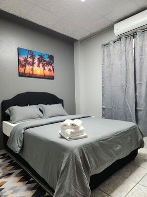 Een net opgemaakt bed met grijze lakens en een opgevouwen handdoek, een schilderij van palmbomen bij zonsondergang aan de muur, en een grijze gordijn.