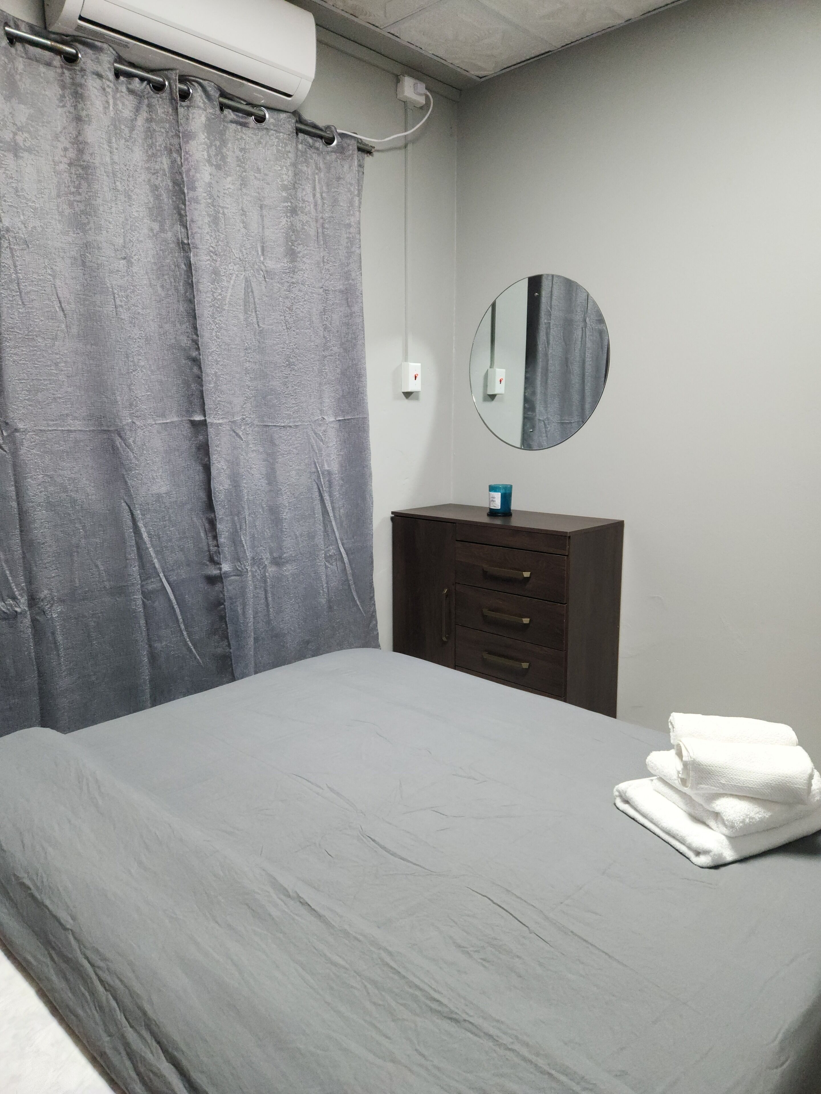 A bedroom with a grey bed, a wooden dresser, a round mirror, and a grey curtain.