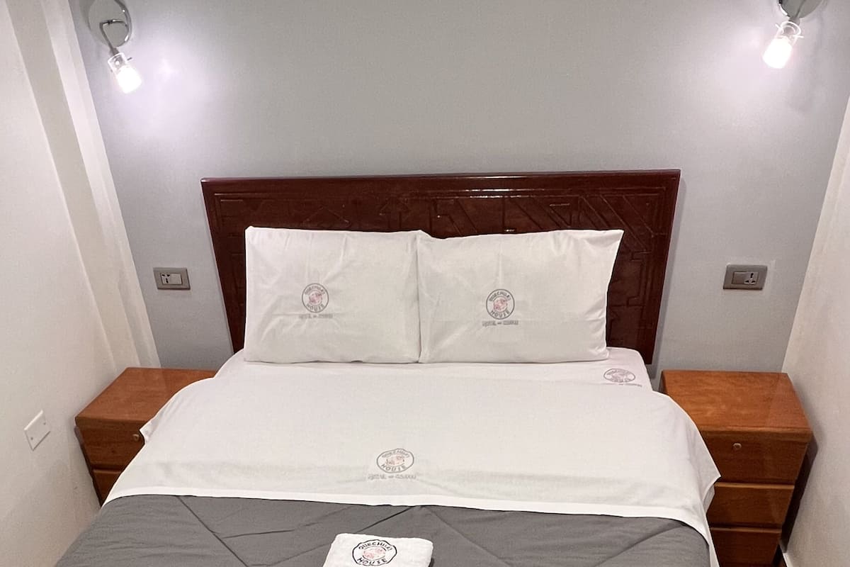 A neatly made bed with a grey quilt and white linens, two bedside tables with lamps, and a wall socket.