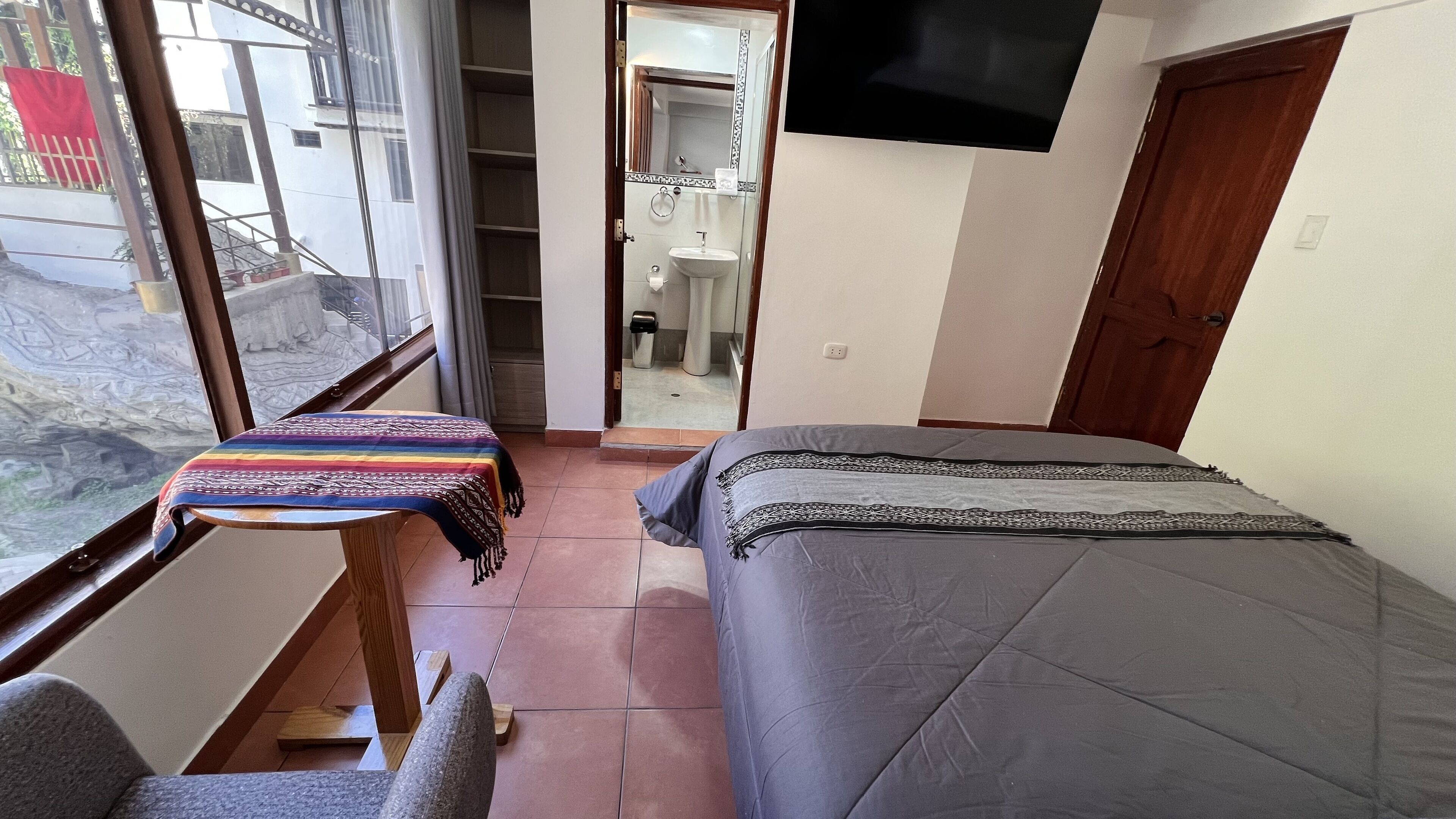 A bedroom with a bed, a small table with a colorful cloth, a TV mounted on the wall, and a view of the outside through a window.