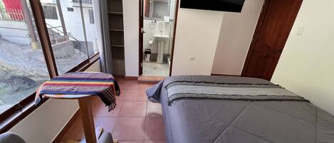 A bedroom with a bed, a small table with a colorful cloth, a TV mounted on the wall, and a view of the outside through a window.