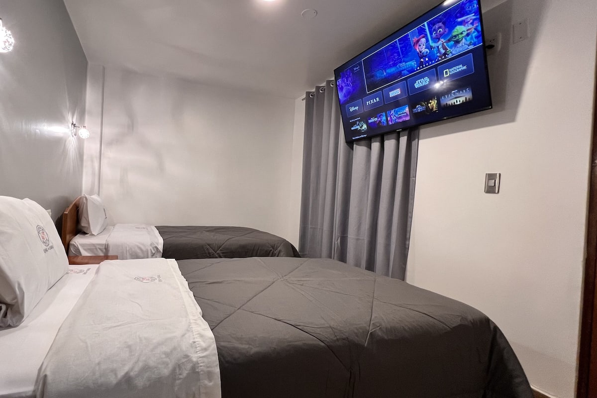 A hotel room with two beds, a flat-screen TV mounted on the wall displaying a movie selection, and a ceiling light fixture.