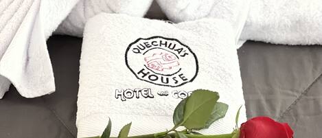A white towel with a heart shape and a rose, and a logo that reads "QUECHUA'S HOUSE HOTEL and GUESTHOUSE".