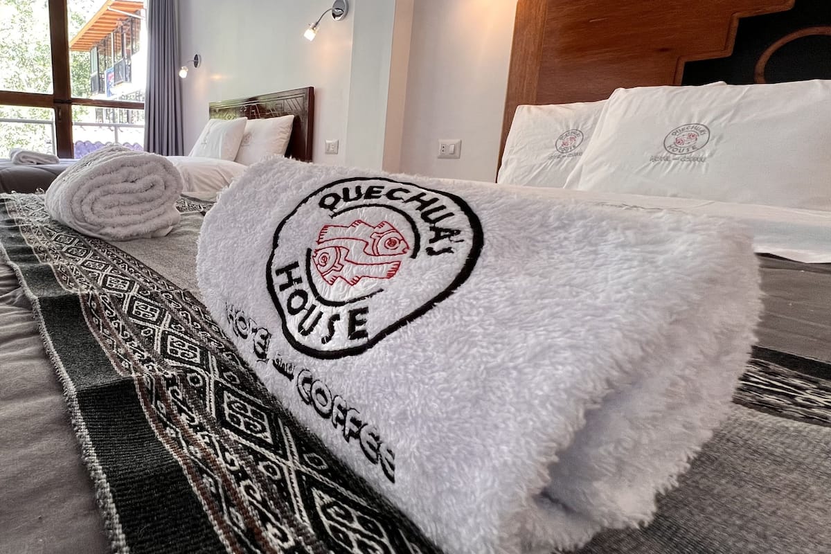 A neatly made bed with a white towel featuring a logo and text, a patterned rug, and a view of a building outside.