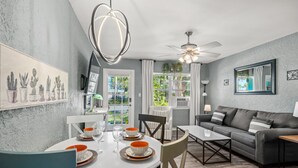 A modern living room with a grey sofa, a dining table with orange and white plates, and a ceiling fan. - Salty Lime Villa 2-Riverview w/pool on riverwalk, near AMI beaches, pet friendly (Bradenton)