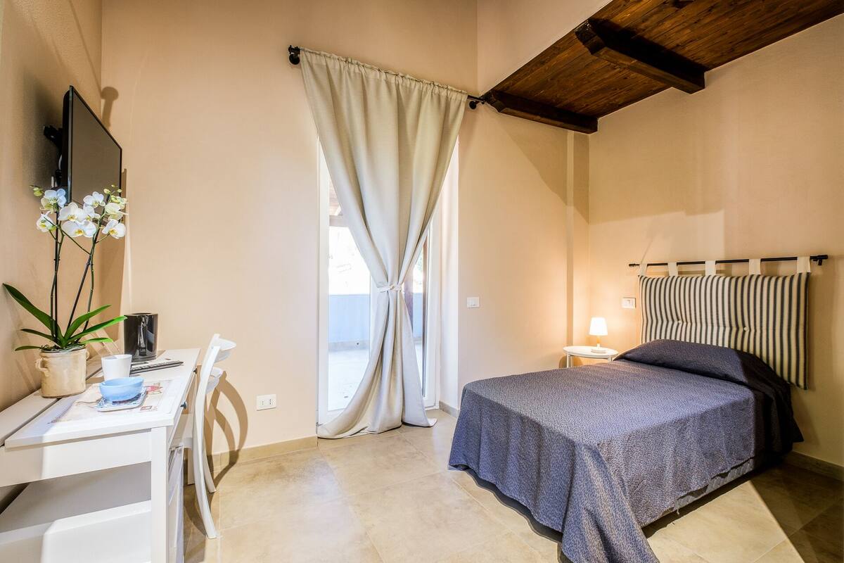 A bedroom with a bed, a desk, a chair, a lamp, and a window with curtains.