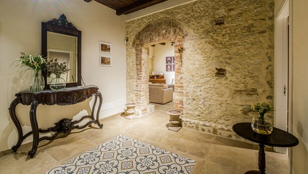 A room with a stone wall, a wooden console table with a mirror, a patterned rug, and a small round table with a vase. - Residenza Mistral (Rende)