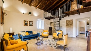 A spacious living room with a blue sofa, yellow armchairs, and a unique animal-shaped coffee table. A wooden staircase with a metal railing leads to an upper level. - Residenza Mistral (Rende)
