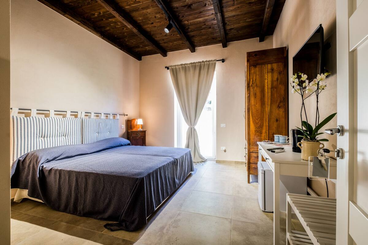 A bedroom with a bed, bedside lamp, curtains, a wooden door, a small table with a plant, and a radiator.