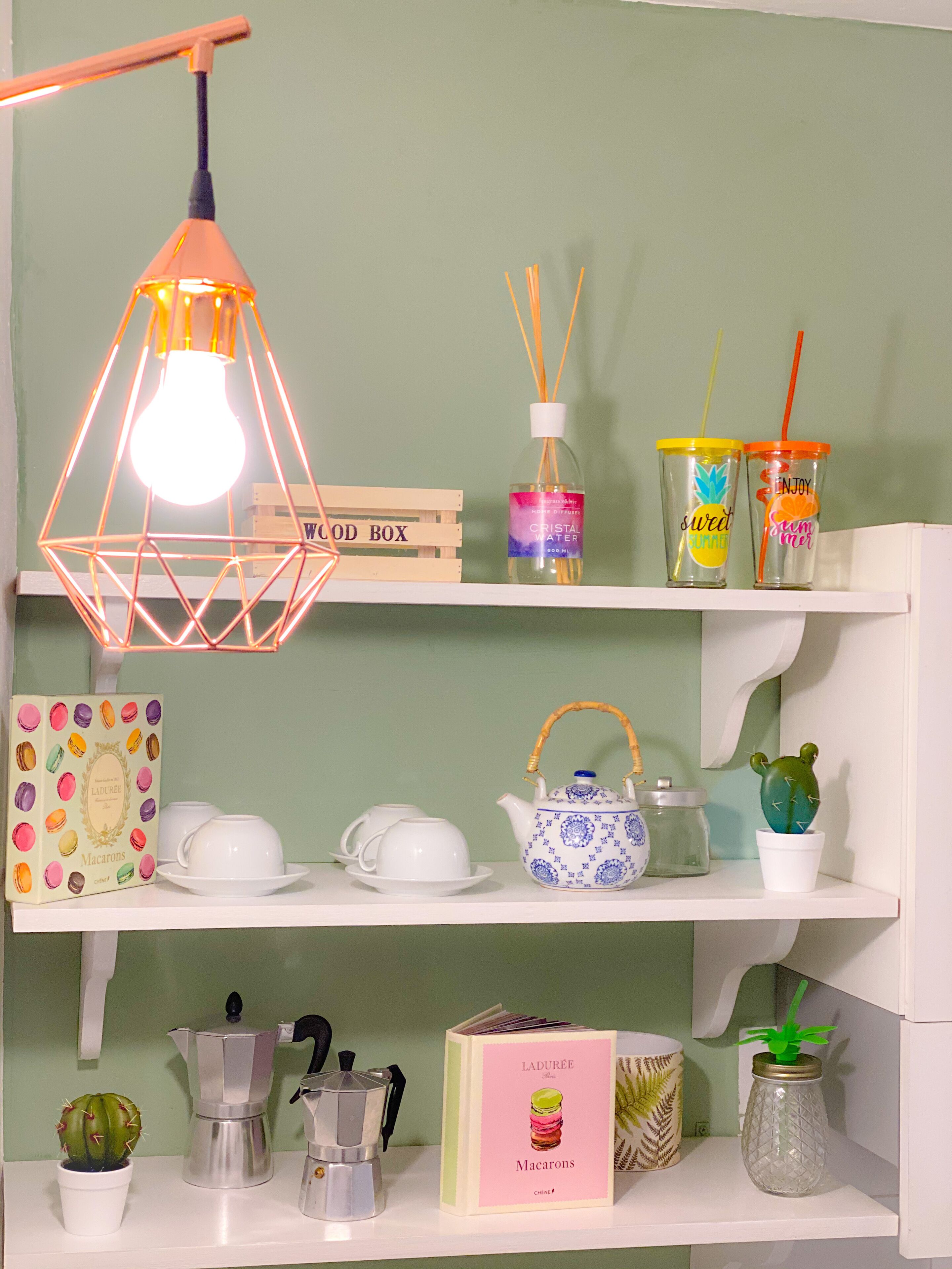 A shelf with a wooden box labeled "WOOD BOX," a bottle of "CRISTAL WATER," two colorful cups with straws, a teapot, a cactus, a macaron box, a coffee maker, and a decorative plant in a glass jar.