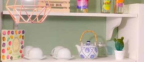 A shelf with a wooden box labeled "WOOD BOX," a bottle of "CRISTAL WATER," two colorful cups with straws, a teapot, a cactus, a macaron box, a coffee maker, and a decorative plant in a glass jar.